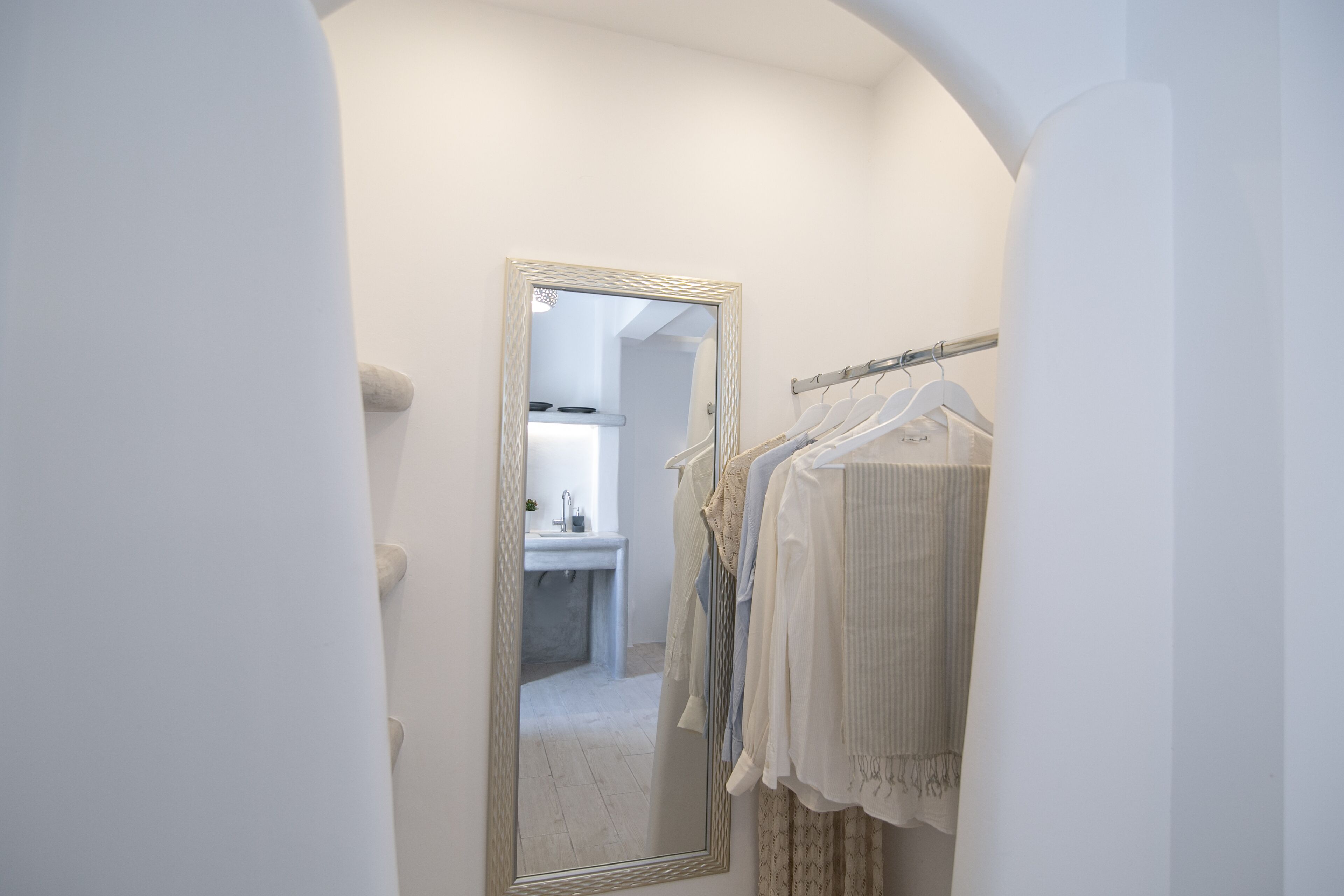 A narrow hallway with a mirror reflecting a bathroom and a clothing rack with hanging garments.