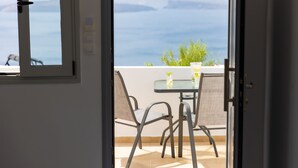 An open door to a balcony with a table and chairs, overlooking a scenic view of the sea and mountains. - Sensia (Santorini)