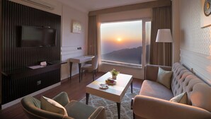 A modern hotel room with a flat-screen TV, a dining area, a sofa, and a large window with a sunset view. - Amritara Surya – A Luxury Spa Retreat (Dharamshala)