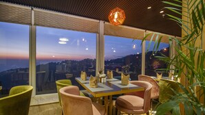 A dining area with a view of the city at dusk, featuring a table with chairs and a hanging light fixture. - Amritara Surya – A Luxury Spa Retreat (Dharamshala)