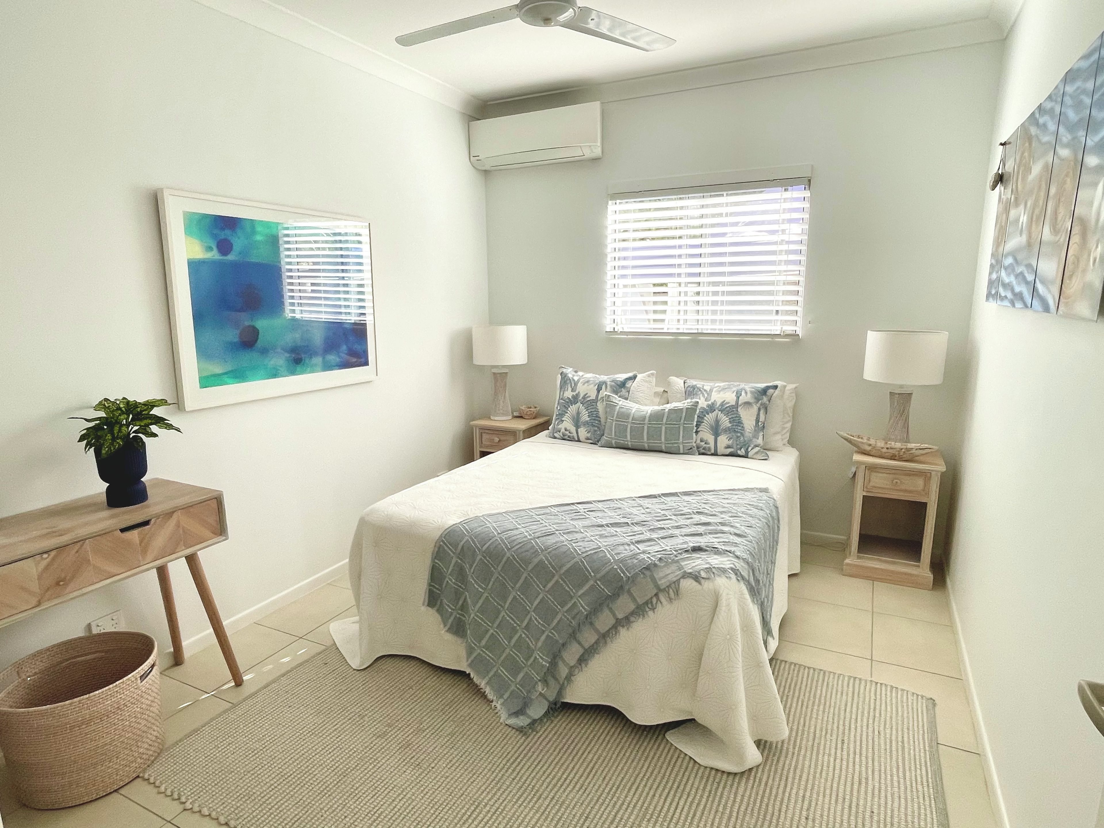 A bedroom with a bed, two bedside tables, a nightstand, a wall-mounted air conditioner, a ceiling fan, a window with blinds, a framed abstract painting, and a potted plant.