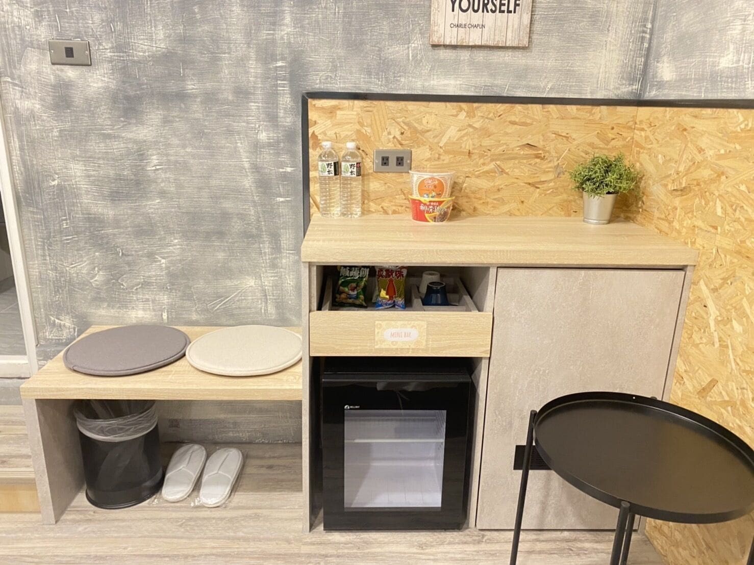 A compact kitchenette with a wooden countertop, a small fridge, a trash bin, and a black chair.