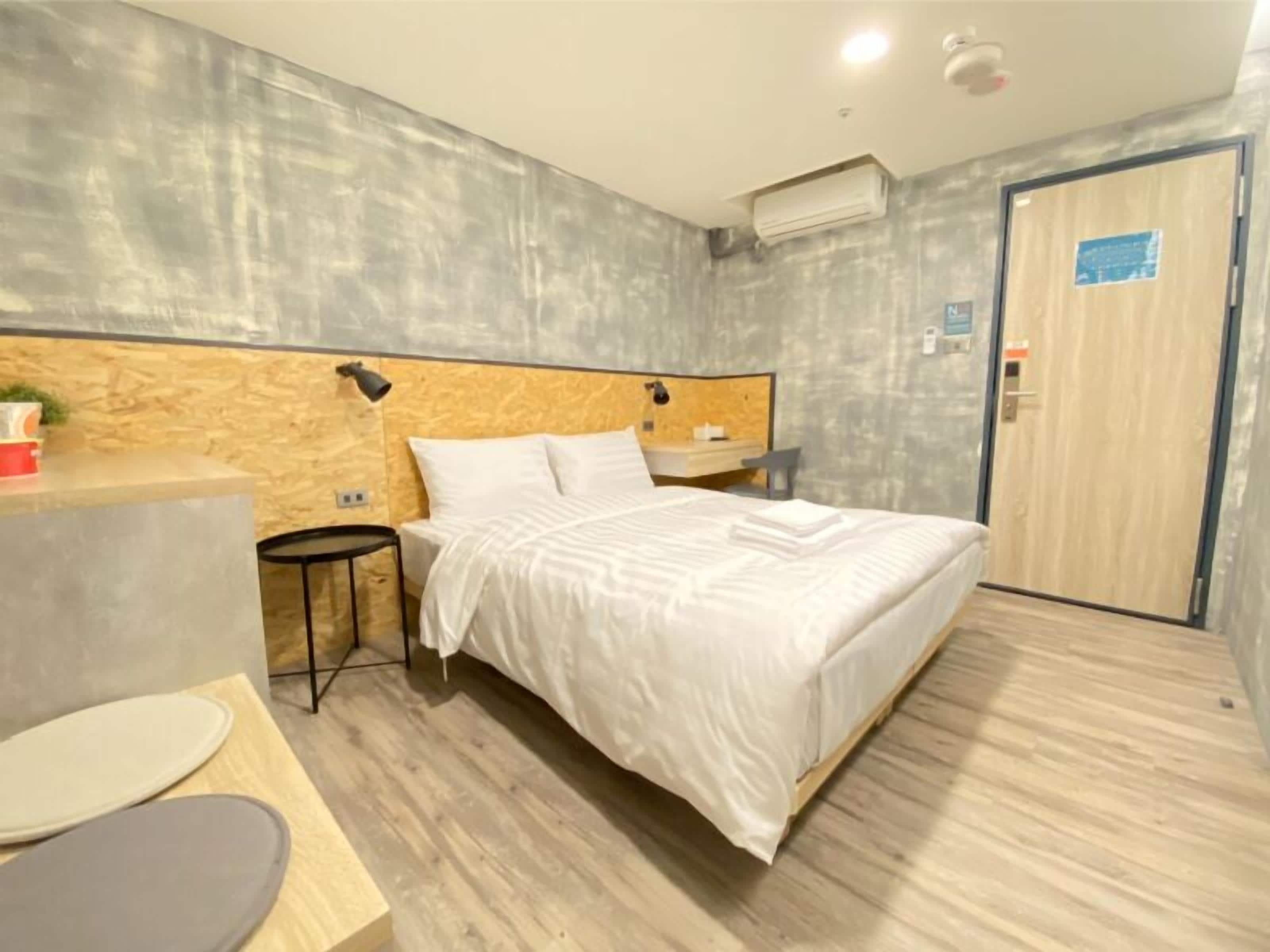 A modern hotel room with a wooden headboard, a bed with white linens, a small bedside table, a desk with a chair, and a door with a sign.