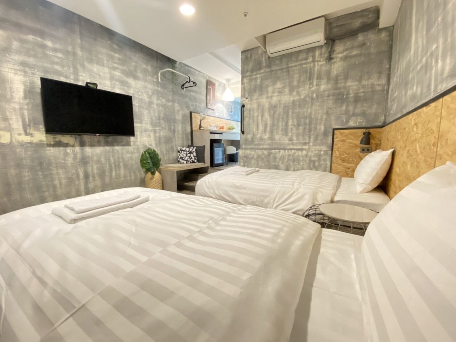 A modern hotel room with two beds, a flat-screen TV mounted on the wall, a small desk with a lamp, and a wall-mounted air conditioner.