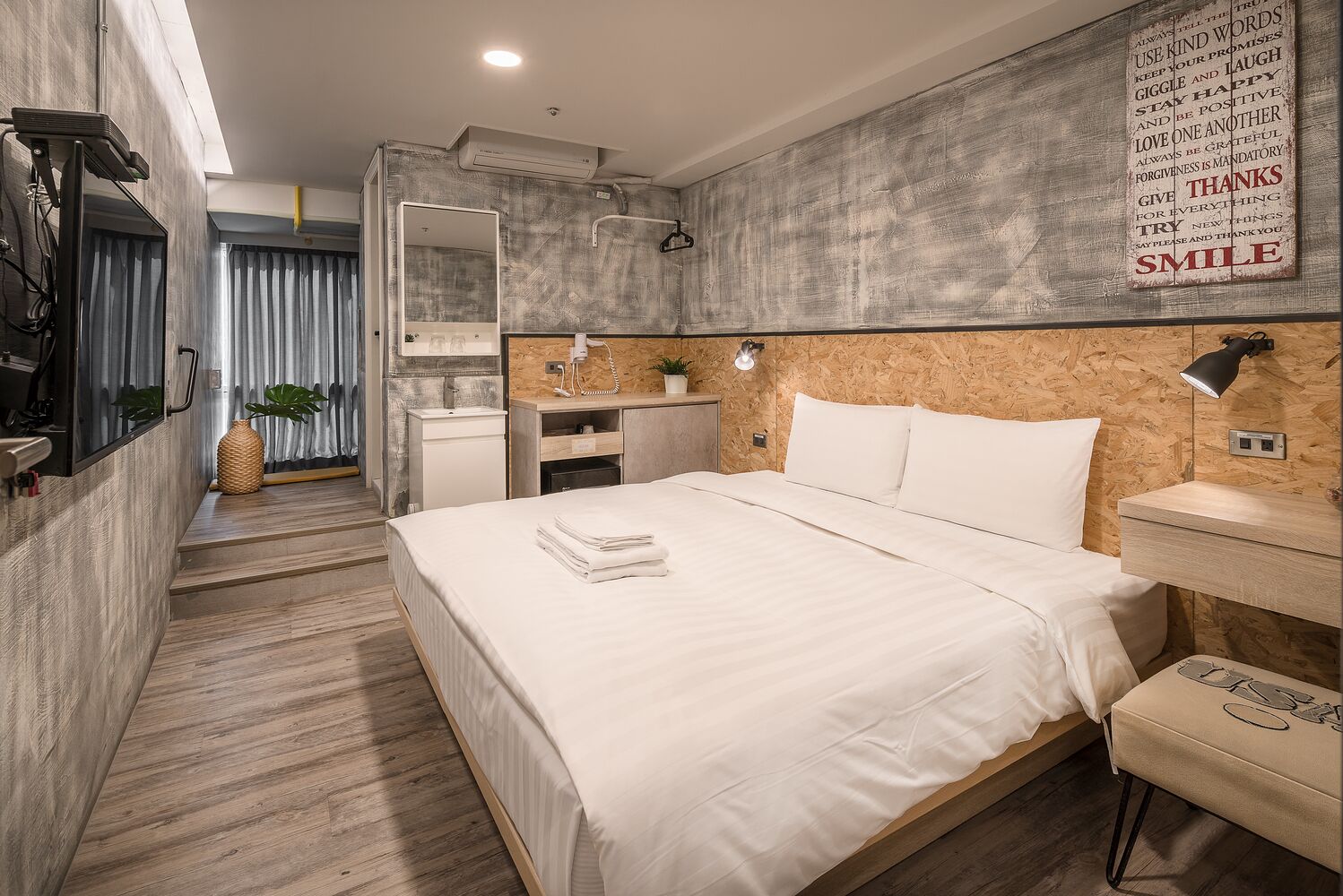 A modern hotel room with a large bed, a vanity with a mirror, a small desk, and a wall-mounted air conditioner.