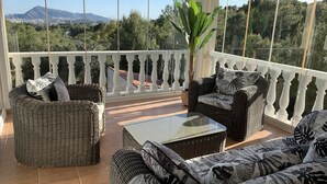 Ein Balkon mit Korbmöbeln, einem Glastisch und Blick auf Berge und Bäume.