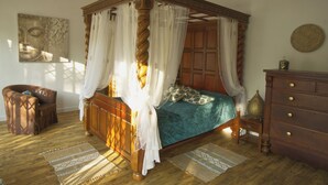 Ein Bett mit Himmelbett, ausgestattet mit einer grünen Bettdecke und weißen Vorhängen, ein Nachttisch aus Holz mit einer Lampe sowie ein brauner Ledersessel.