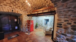 A stone-walled room with a wooden door, a red refrigerator, a ladder, and a small seating area with a chair and a table. - Bel Appartement en sud Ardèche au Bord de la Rivière (Ucel)