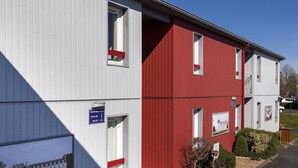 A two-story building with a red and white facade, featuring multiple windows and a small garden area with a bench.