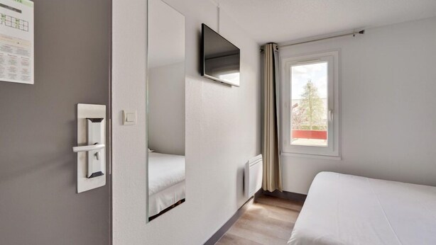 A hotel room with a bed, a mounted TV, a window with curtains, and a wall-mounted thermostat.