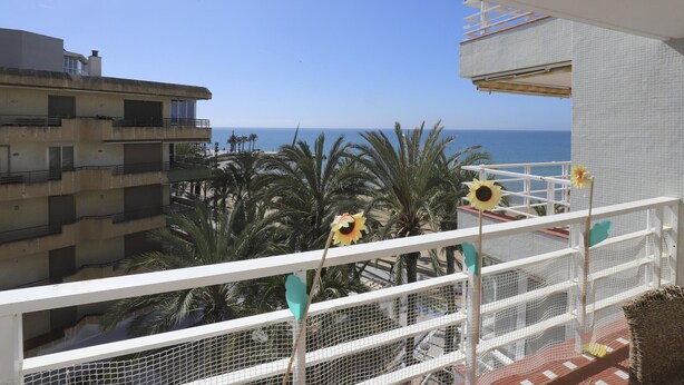 A balcony with a view of the sea, palm trees, and a building with balconies.