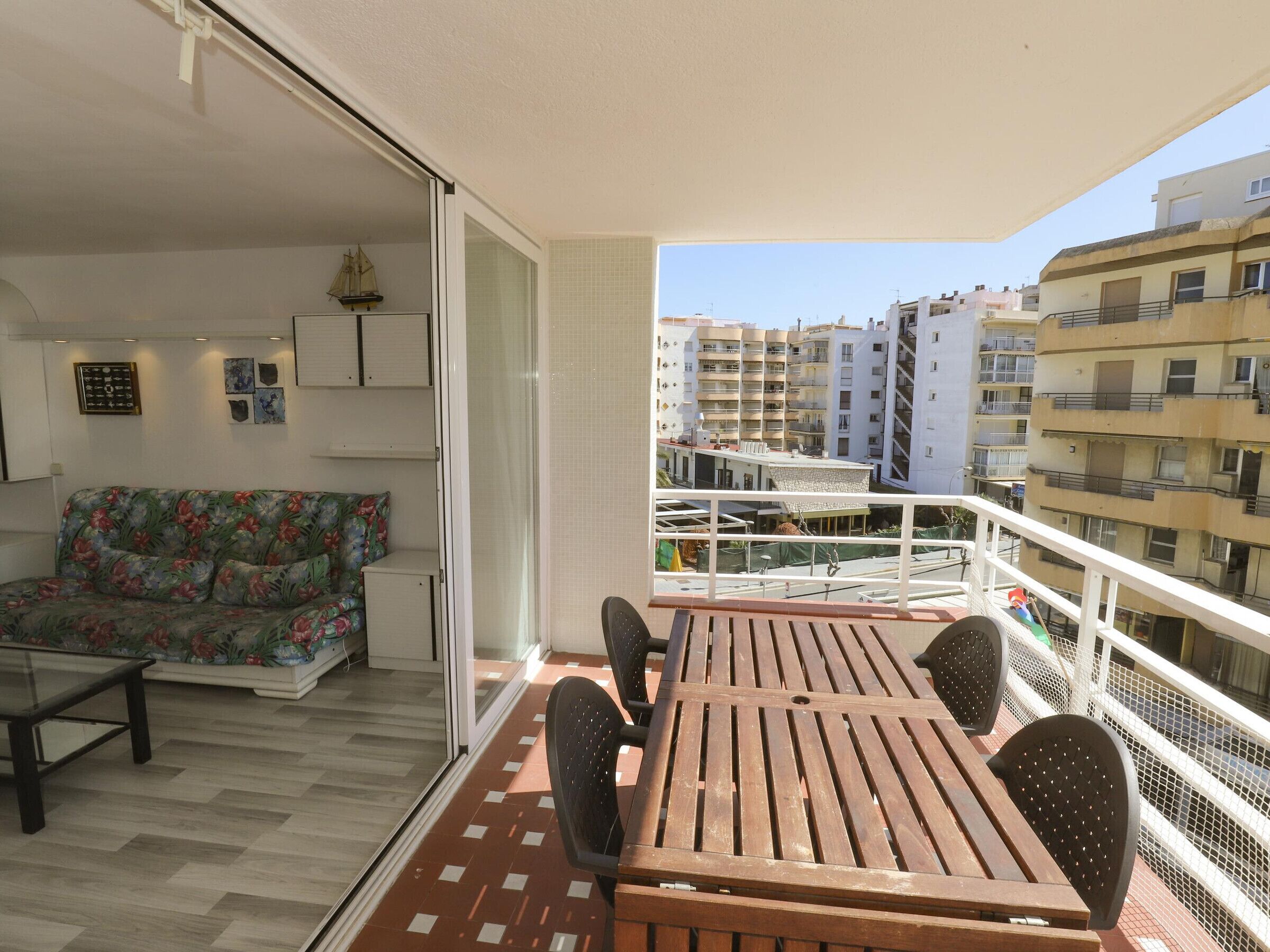 A balcony with a wooden table and chairs, a sofa with floral upholstery, and a view of surrounding buildings.