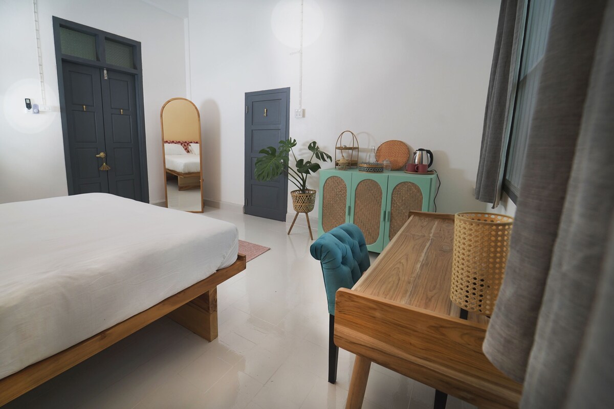 Une chambre à coucher avec un lit, une table en bois, un miroir, une commode de couleur turquoise et une plante.