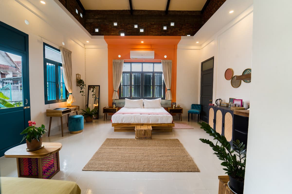 A modern bedroom with a large bed, a wooden headboard, a small table with a plant, a bedside table with a lamp, and a view of the outside through a window.