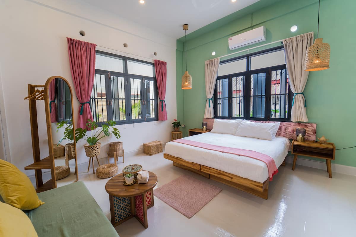 A well-lit bedroom with a large bed, two windows with curtains, a bedside table with a lamp, and a small round table with a plant.