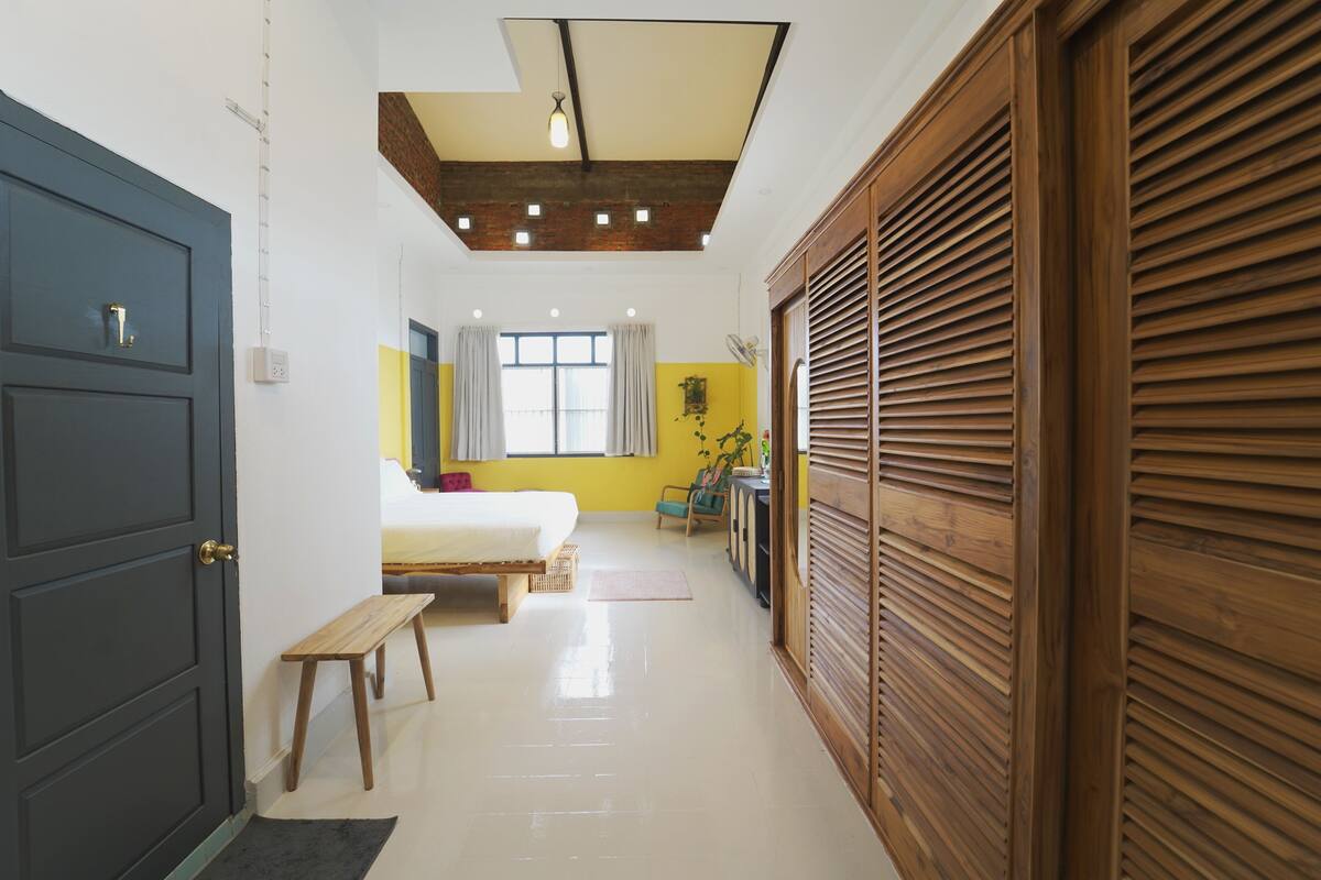 A hotel room with a bed, a bench, a wooden wardrobe, a ceiling fan, and a window with curtains.