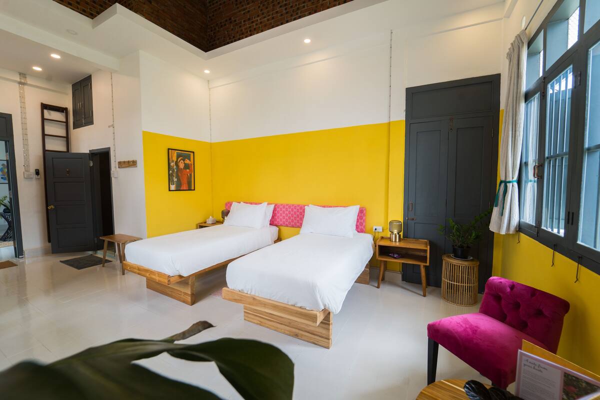 A hotel room with two beds, a wooden floor, a bright yellow accent wall, a large window with curtains, and a pink chair.
