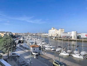 Un puerto deportivo con barcos amarrados, una carretera con coches y bicicletas, y edificios al fondo.