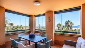 Ein Balkon mit Tisch und Stühlen, einer Couch und großen Fenstern mit Blick auf den Strand und Palmen.