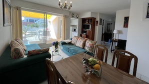 Una sala de estar con un sofá, una mesa de comedor con sillas, una araña y vistas a un área exterior a través de un ventanal corredero.