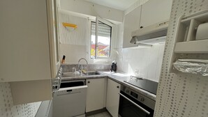 Cocina compacta con armarios blancos, lavavajillas, estufa y ventana con persianas.