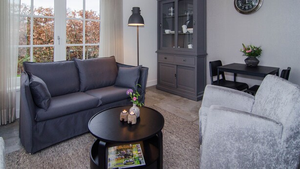 A living room with a grey sofa, a round coffee table with magazines, a cabinet, and a dining table with chairs. - Rural B&B with a beautiful outdoor view and a village atmosphere! (Rhenen)
