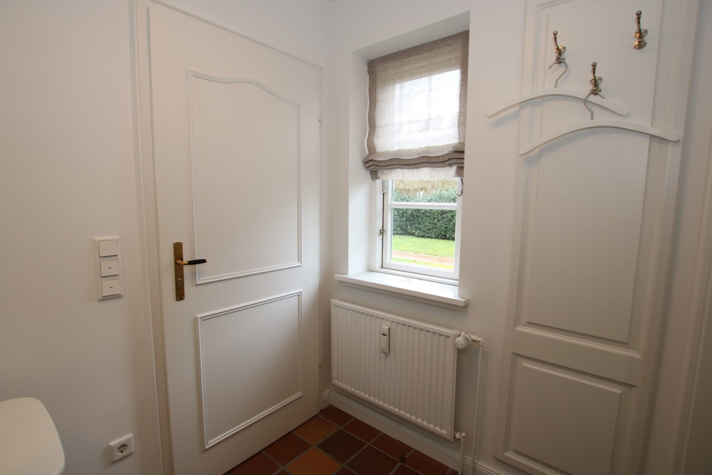Ein Zimmer mit weißer Tür, einem Fenster mit Vorhang, einem Heizkörper und einem an der Wand befestigten Kleiderhaken.