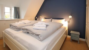 A neatly made bed with folded towels, a bedside table with a lamp, and a window with curtains. - Ferienwohnung in Wrixum, mit 3- Zimmern auf 60m² im Reetdachhaus (Wrixum)