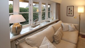 A cozy room with a beige sofa, a window with a view of greenery, a lamp, and a framed picture on the wall. - Ferienwohnung in Wrixum, mit 3- Zimmern auf 60m² im Reetdachhaus (Wrixum)