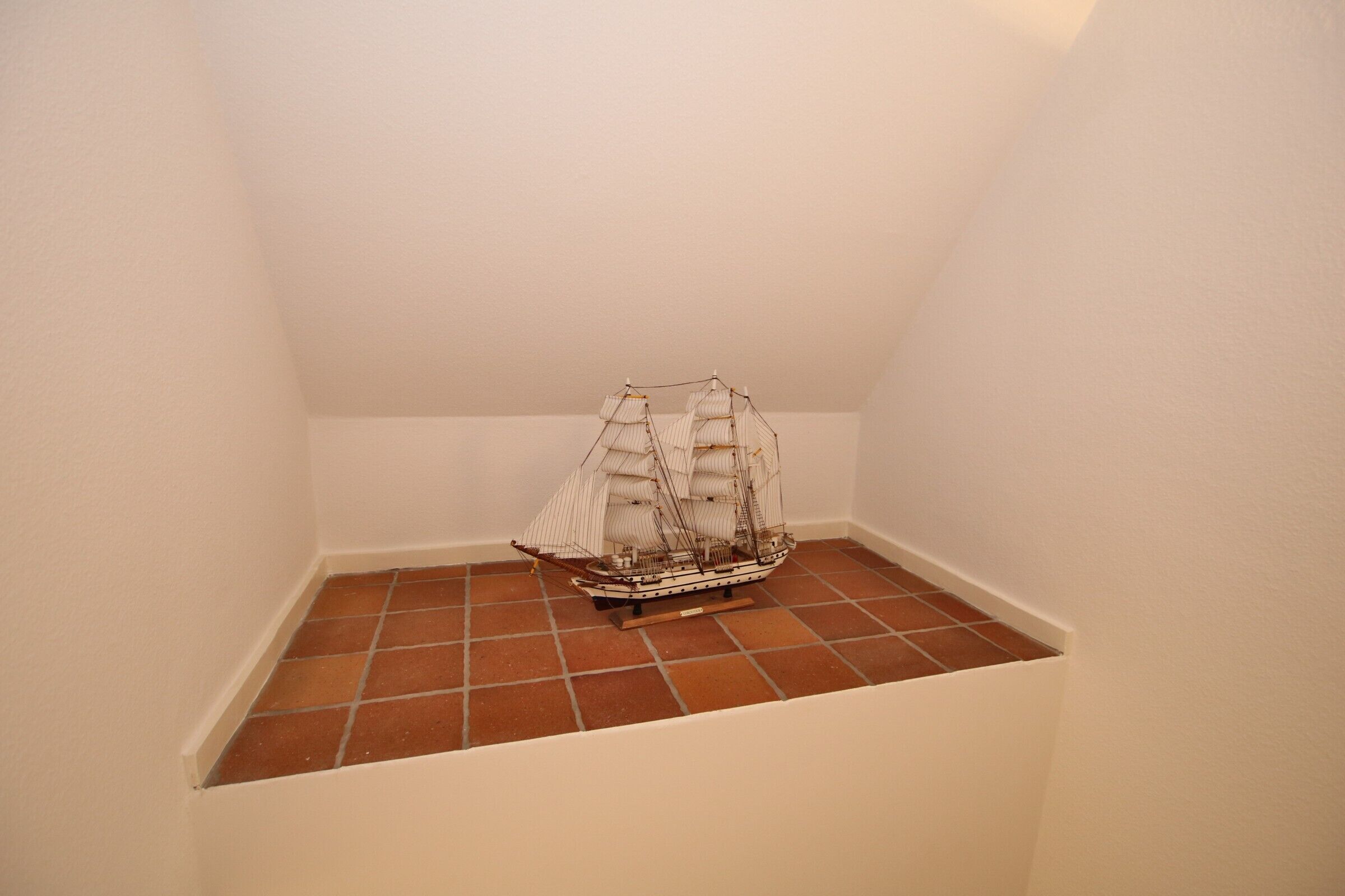 A model ship in a corner of a room with white walls and terracotta floor tiles.
