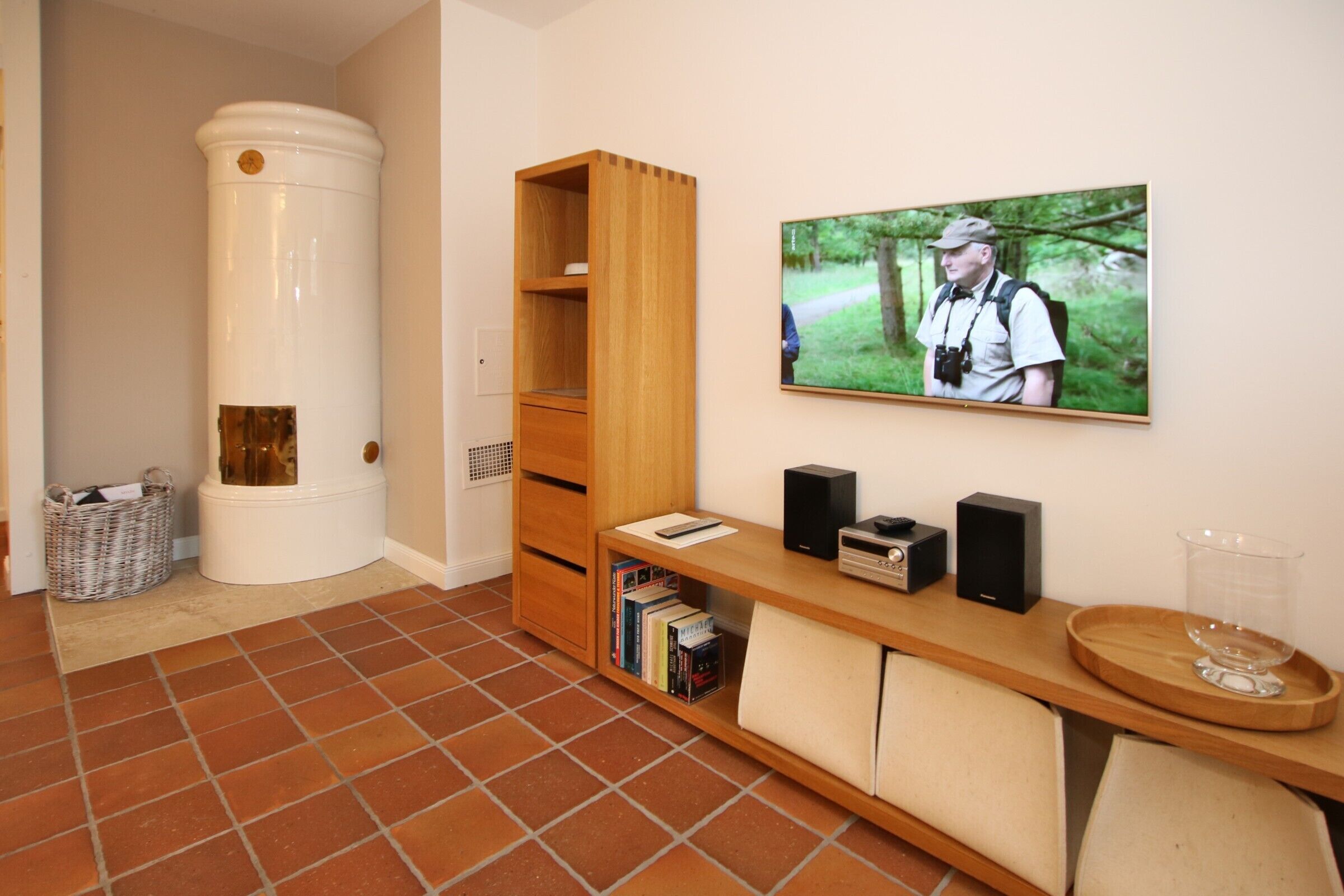 A room with a white toilet, a wooden cabinet, a television mounted on the wall, a bookshelf, and a tray with a glass.