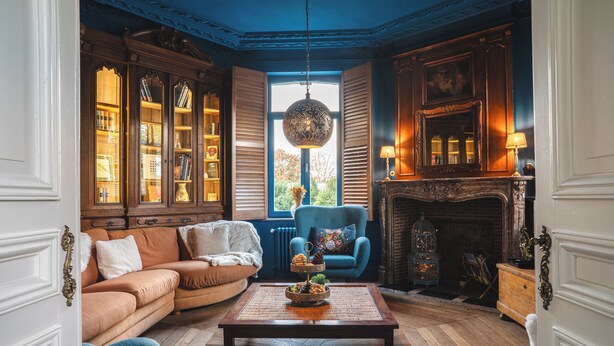 Una habitación tradicional con piso de madera, un sofá, un sillón azul, una chimenea y un ventanal con contraventanas.