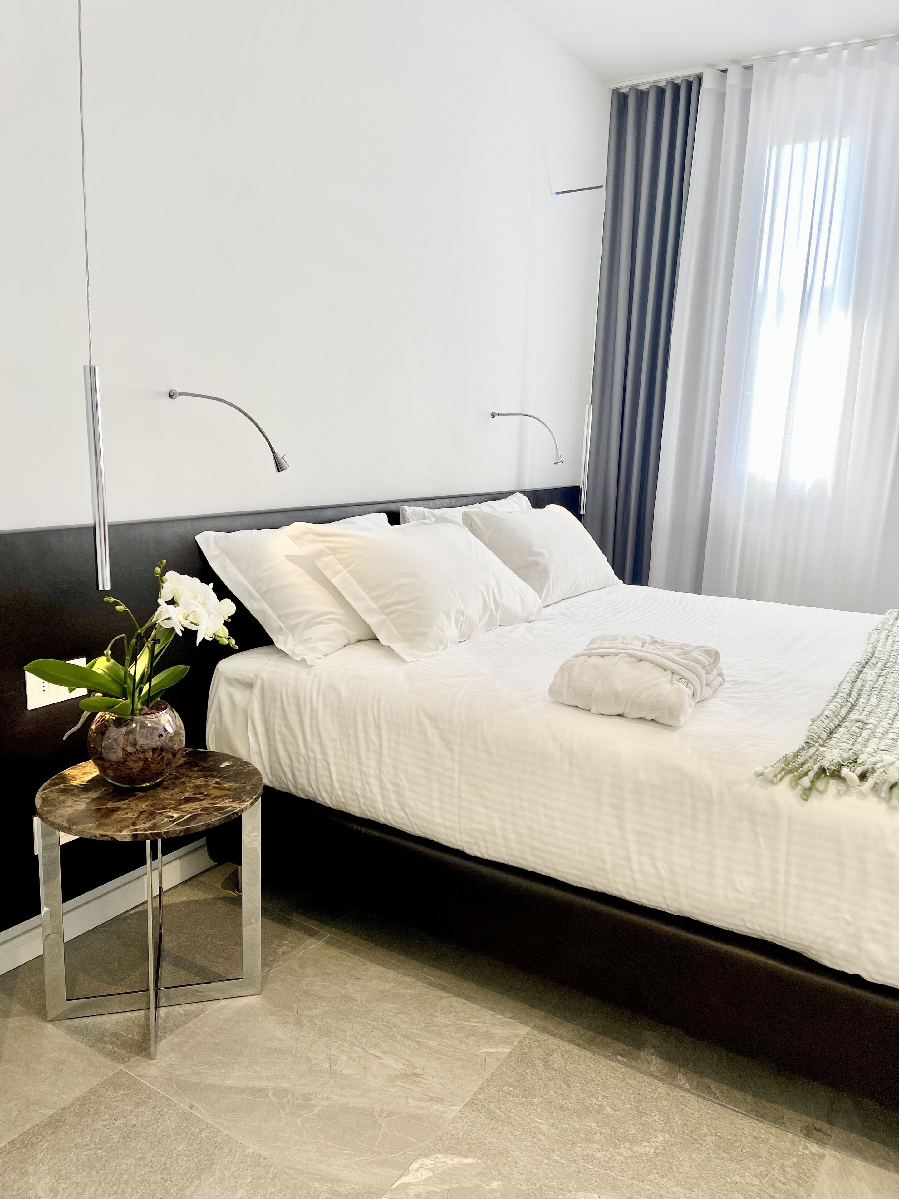 Een moderne slaapkamer met een bed, witte beddengoed, een klein tafeltje met een bloevas en een nachtkastje.