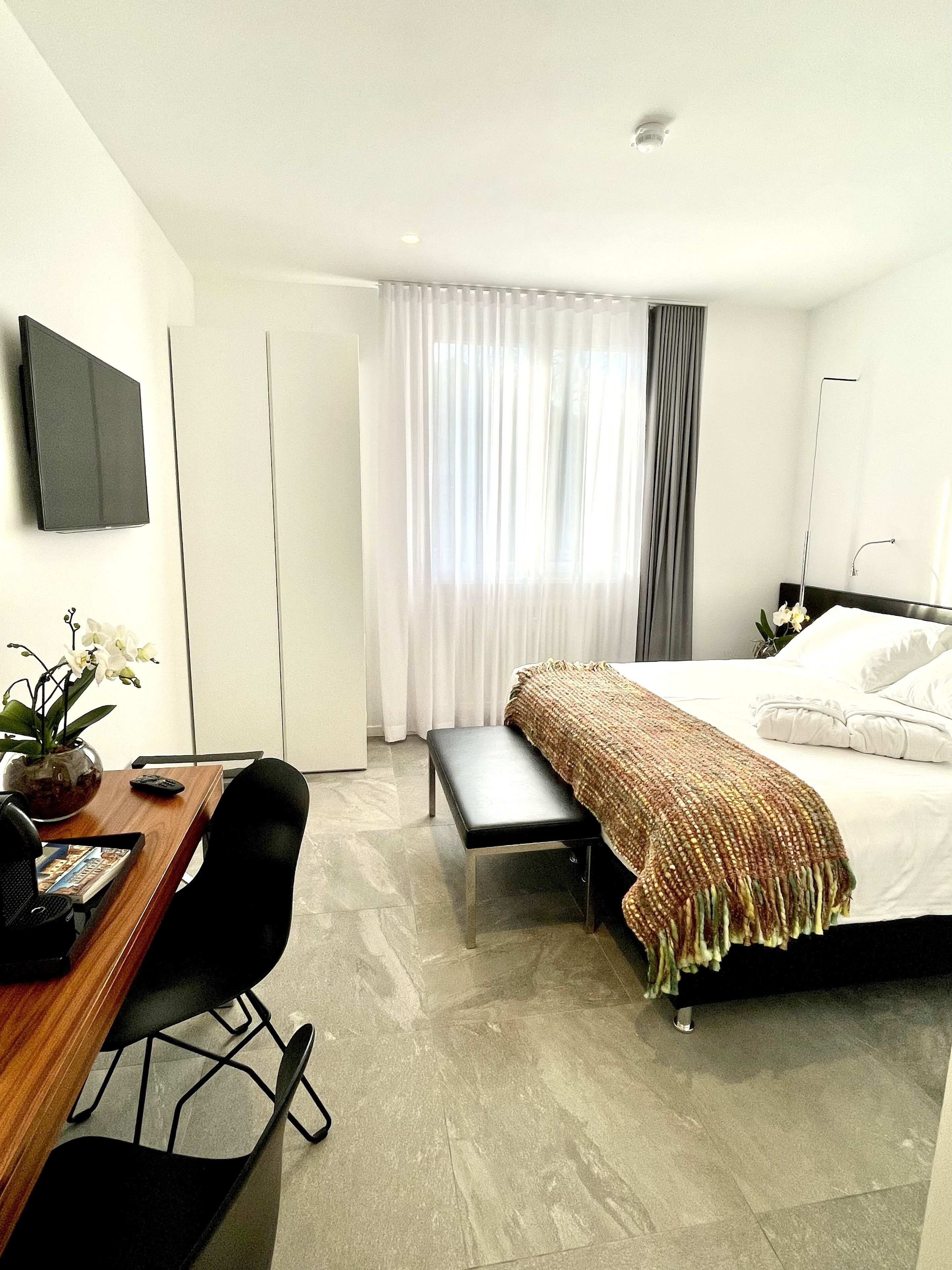 A modern hotel room with a bed, a desk with a chair, a small table, a potted plant, and a large window with curtains.