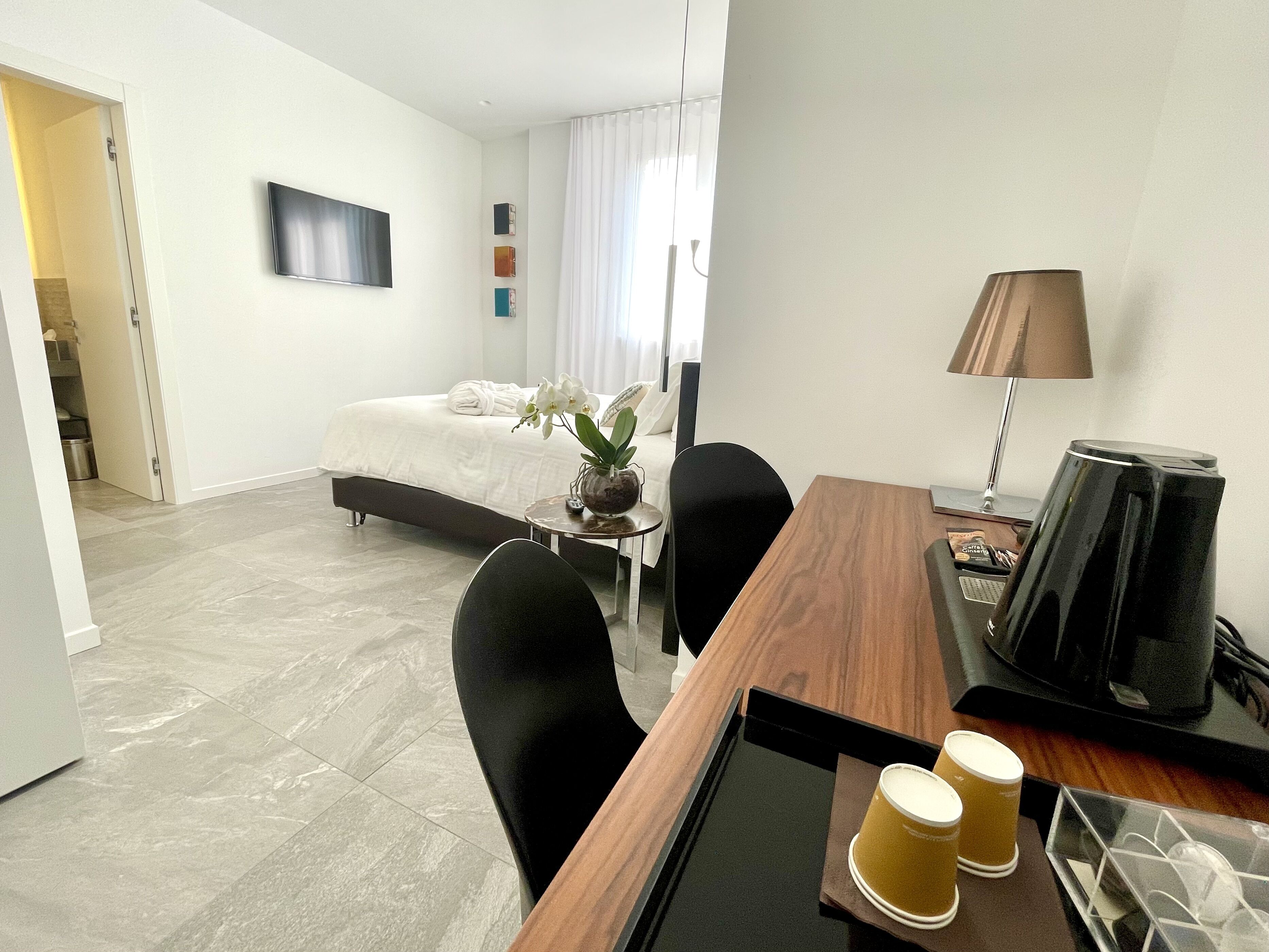 A hotel room with a bed, a desk with a lamp, a coffee maker, and a small table with two cups.