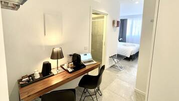 A hotel room with a wooden desk, a laptop, a coffee maker, and a chair. There is a bed with a white comforter and a window with curtains.