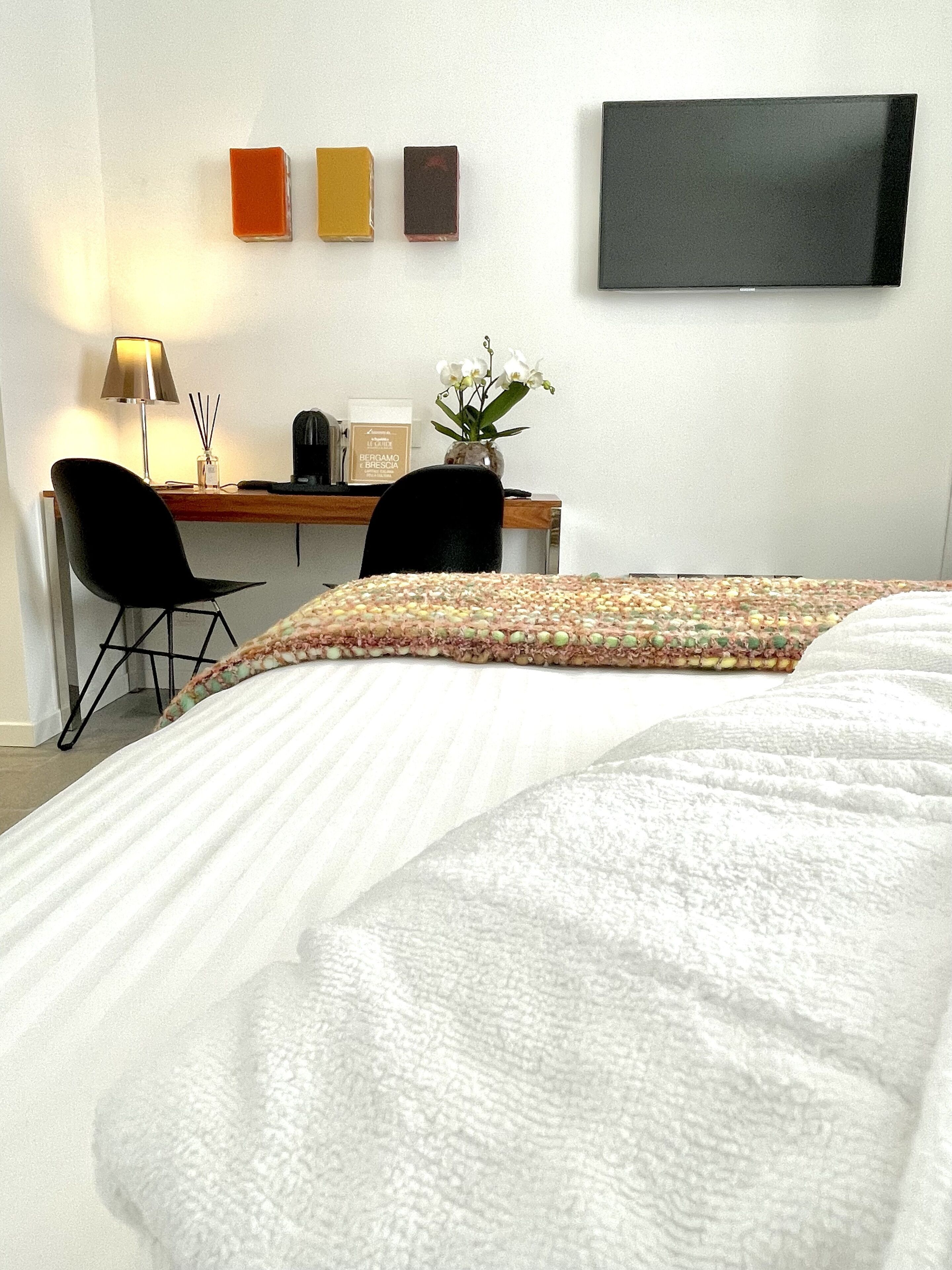 Ein Hotelzimmer mit Bett, zwei Sesseln, einem Schreibtisch mit Computer und einem Fernseher an der Wand, sowie einer kleinen Topfpflanze.