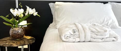 A neatly made bed with a folded towel, a vase with white flowers, and a modern shower head.