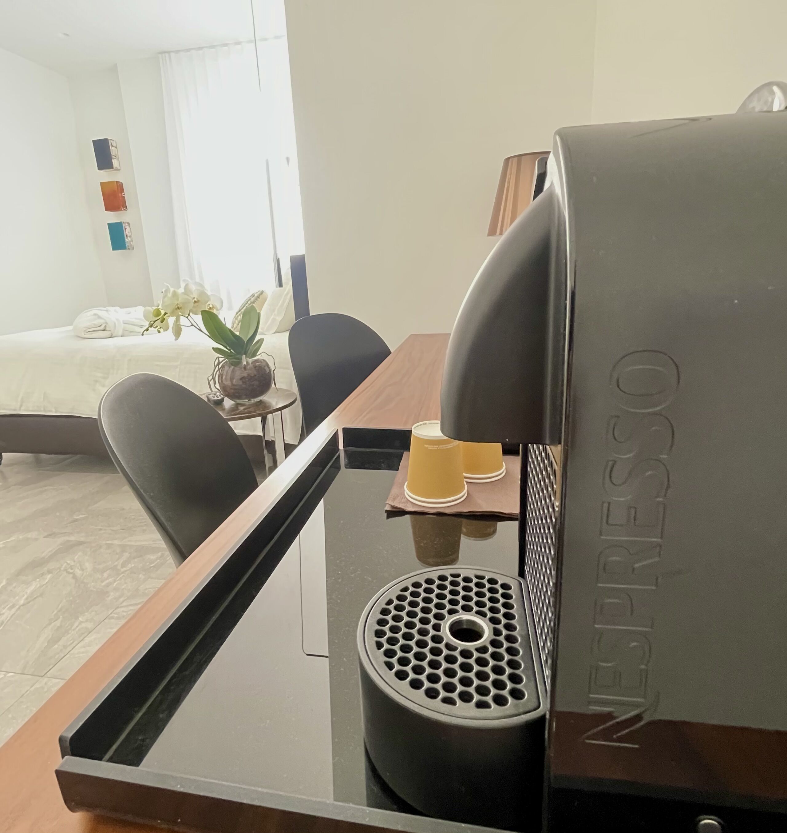 Een hotelkamer met een bed, een bureau met een koffieapparaat, een stoel en een kleine tafel met een plant en een kop.