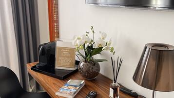Hotelkamer met een houten bureau, een laptop, een afstandsbediening, een boek, een vaas met witte bloemen, een lamp en een aan de muur bevestigde televisie.