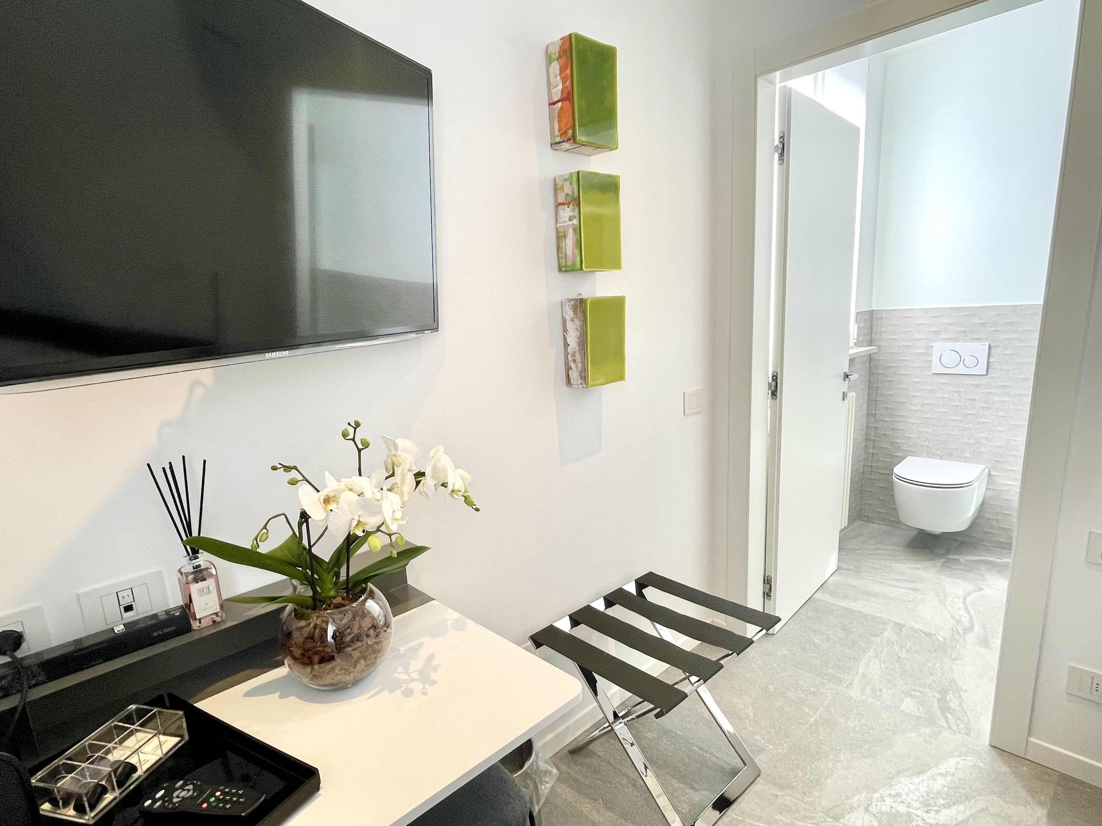 A modern bathroom with a toilet, a shower, and a sink. There is a TV mounted on the wall and a vase with flowers on the counter.