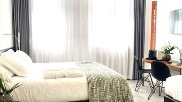 Habitació d'hotel moderna amb un llit, un escriptori i una cadira. Hi ha una finestra gran amb cortines, una televisió de pantalla plana i un vas amb flors.