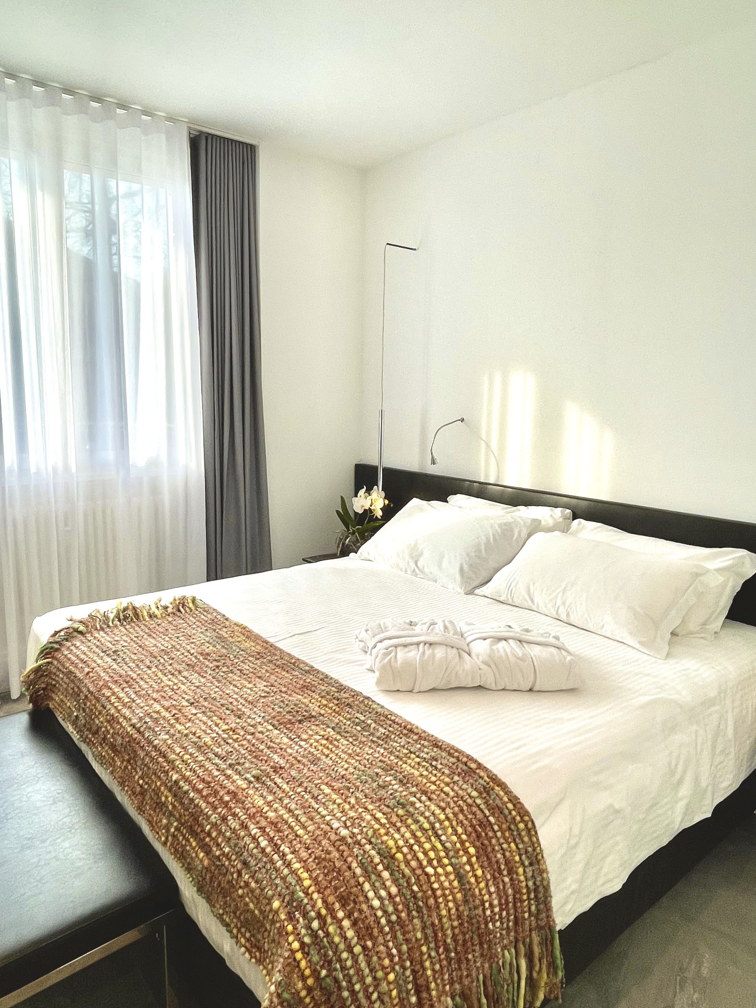 A neatly made bed with white linens, a brown woven throw blanket, and a bedside table with a vase of flowers.