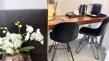 A modern hotel room with a wooden desk, two black chairs, a coffee maker, and a vase with white orchids.