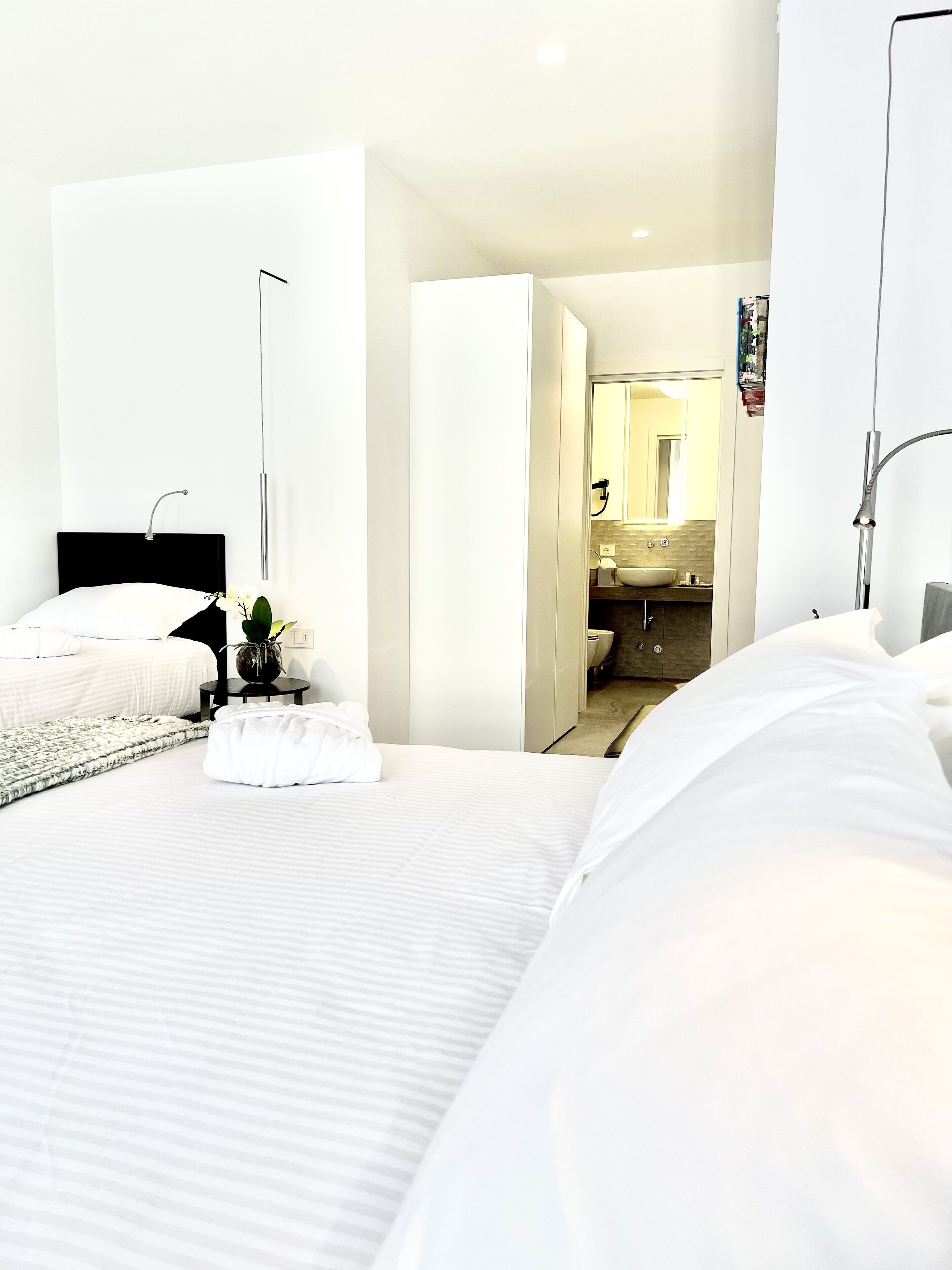 A modern hotel room with a large bed, a bedside table with a lamp, a small plant, and a bathroom visible through an open door.
