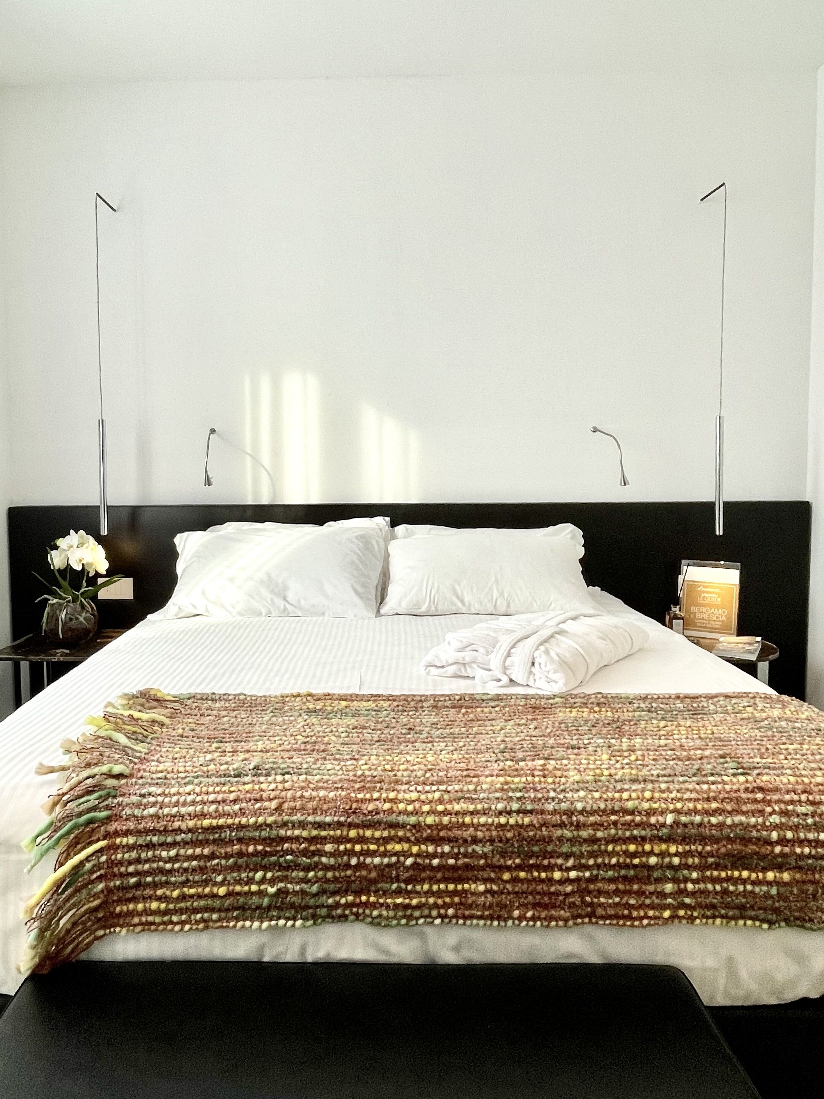 Un letto rifatto con lenzuola bianche, una coperta colorata in tessuto e una panca nera ai piedi del letto.
