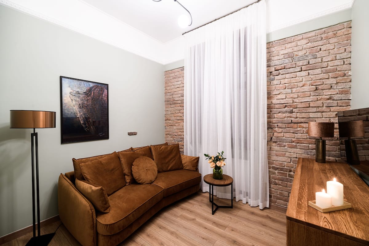 Un salon moderne avec un canapé marron, une petite table ronde surmontée d’un vase, un mur en briques et un plancher en bois.