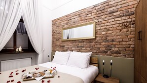 Slaapkamer met een bakstenen muur, een bed met witte lakens, een nachtkastje met kaarsen, en een dienblad met gebak en koffie.