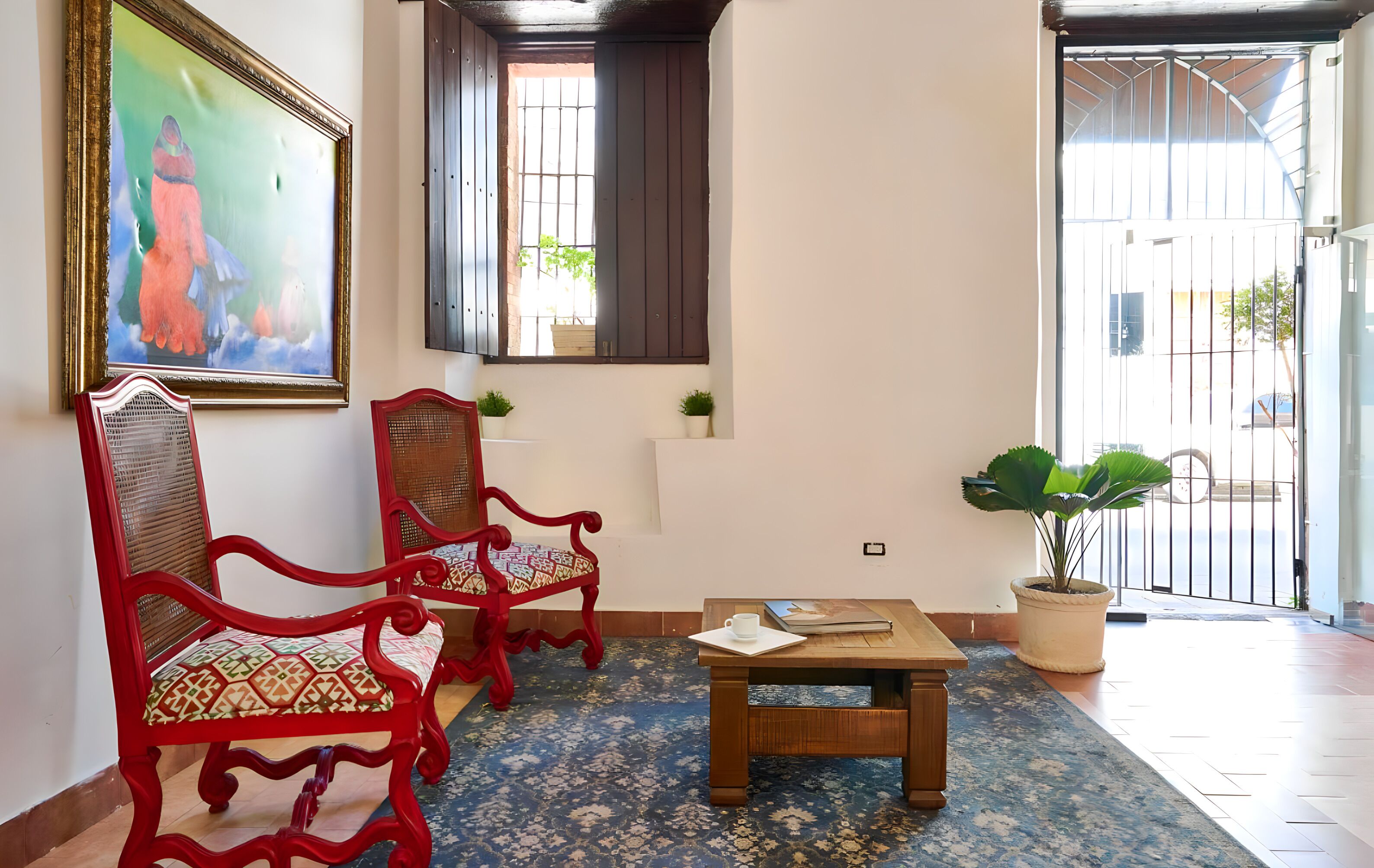 Een kamer met twee rode stoelen, een klein houten tafeltje met een kopje en notitieboekje, een groot schilderij, een raam met zonwering en een kamerplant.