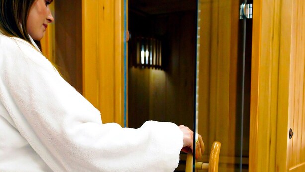 A person in a white robe opening a wooden cabinet with a glass door. - Klemi (Trento)
